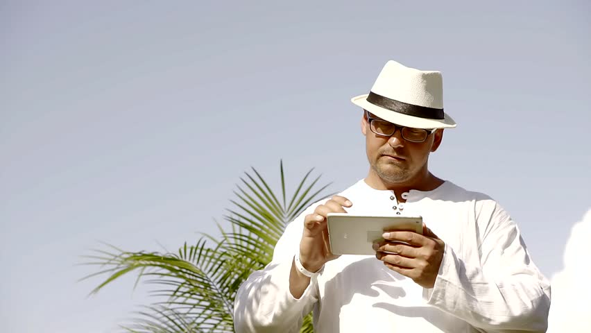 Handsome man is going through the newsfeed in social network on his tablet during vacation on an island. He is wearing light white longsleeve, white hat and glasses.
