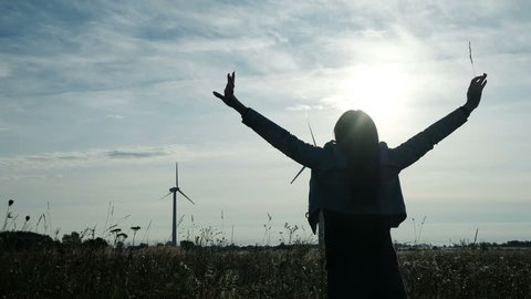 One free happiness young woman joyful turn with opening raised hands on the grassland. The spring weather is natural sunny and windy, at skyline in horizon clouds. Background eco electrical windfarm