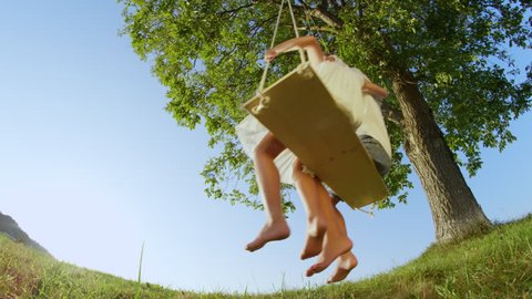 SLOW MOTION LOW ANGLE: Cheerful couple of kids embracing on big swing. Happy little brother and sister have fun swaying on swing in sunny spring. Smiling boy and girl hugging and swinging under a tree