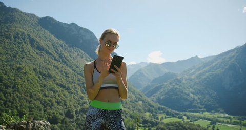 Fit Young Woman in Sportswear Uses Touchscreen Smartphone. She Wears Sunglasses, in the Background Breathtaking Hills, Mountains, and Valley View.  Shot on RED Epic 4K UHD Camera.