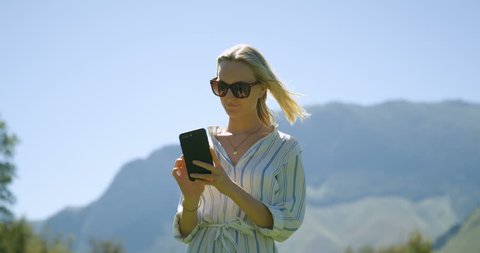 Beautiful Young Blonde Woman in the Casual Clothes and Sunglasses Uses Mobile Phone while Standing on the Meadow with Hills and Mountains in the Background. Shot on RED Epic 4K UHD Camera.
