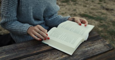 Close-up of the Woman's Hands while She Reads a Book. She Sits Outside, Wears Knitted Sweater and Holds the Book on the Wooden Table. Shot on RED Epic 4K UHD Camera.