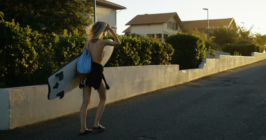 Following Shot of the Beautiful Young Woman Holding Surfboard and Walking Through Suburban Area Towards the Beach. She Has Blond Hair and Sun is Shining. Slow Motion. Shot on RED Epic 4K UHD Camera.
