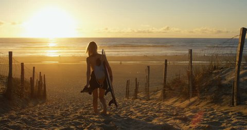 Side View Footage of Beautiful Young Woman Carrying Surfboard Walking Towards the Sea. Girl Has Blonde Hair and a Backpack.The Beach is Empty and Sun is Setting.  Shot on RED Epic 4K UHD Camera. स्टॉक वीडियो