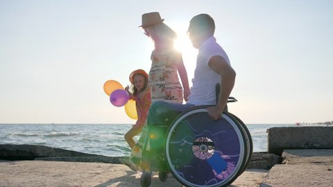 kid at holiday on Sea together with disabled parents, invalid in wheelchair walks with wife and daughter