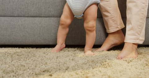 Close up of baby's feets making his first steps with mom on the carpet and coming closer to the camera at home. Inside