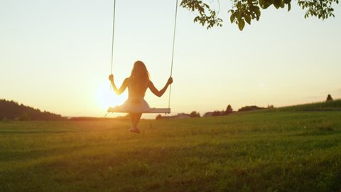 SLOW MOTION LENS FLARE: Smiling young woman swaying on rope swing under big tree at sunrise. Pretty girl in white dress swinging over the sun at golden sunset. Beautiful lady enjoying summer evening