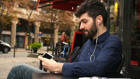 Handsome young man making video call to girlfriend by smartphone using earphones. Attractive male person wears blue shirt with grey jeans and sitting at street cafe. Concept of using mobile phone