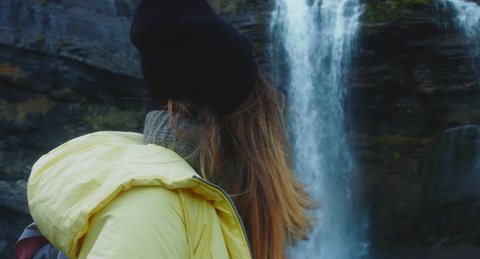 CU portrait of young Caucasian female hiker in yellow raincoat wearing backpack enjoys the view of a beautiful waterfall in French Alps. 4K UHD 60 FPS SLO MO  – Video có sẵn