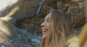 SMARTPHONE CAMERA POV Caucasian female hiker with a backpack making a selfie or video call near small waterfall in the mountains. 4K UHD 60 FPS SLO MO