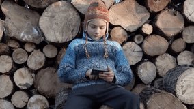 The boy is playing on the smartphone and hiding it behind his back. Portrait of a child on the background of firewood playing on a smartphone. 