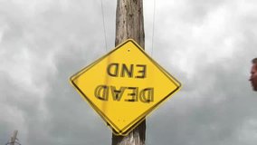 Yellow dead end sign swings from side to side and is upside down, end of clip stays still, in natural sunlight.