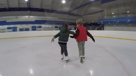 Small girl and boy ride together on ice skates holding hands on covered ice rink.