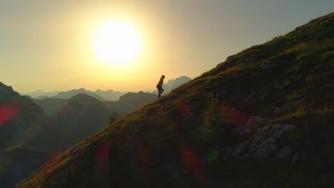 AERIAL SILHOUETTE: Camera flying along a contour of young woman hiker walking up a grassy, rock-specked hill at golden sundown. Female hiker ascending a mountaintop in stunning amber summer evening.