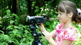 Young girl looks into video camera on background of green park background. Children outdoors in summer are creative work of cinema. Beautiful footage for family. Interesting world in childhood.