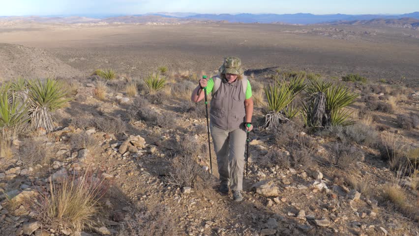 Hiking women in sun protective clothing with a backpack and trekking sticks, it rises along the path up the hill in the Mojave desert. Front view, slow motion. National Park Joshua Tree, USA.
