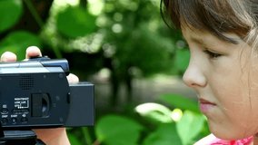 Young girl looks into video camera on background of green park background. Children outdoors in summer are creative work of cinema. Beautiful footage for family. Interesting world in childhood.
