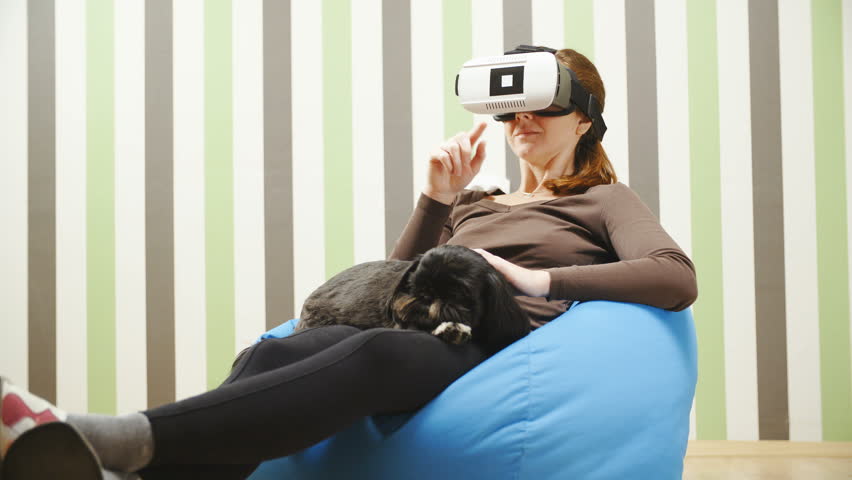 Person surfing the web with VR glasses while caressing dog 4K. Long shot wide view of person in focus sitting on bean bag using virtual reality space for web browsing. Pet on lap resting.