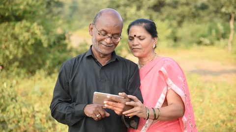Happy senior Indian couple husband and wife using smartphone, looking at  photos on smartphone, smiling and laughing together at outdoor. 