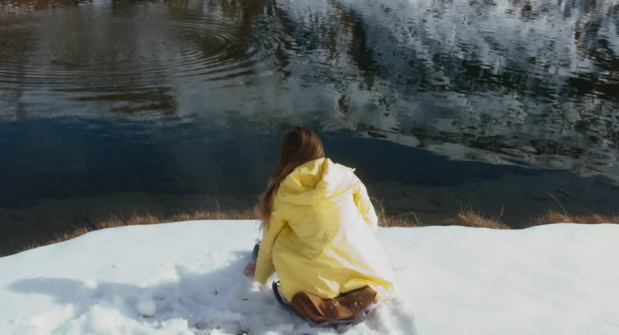 Young female hiker with backpack halts near peaceful mountain lake, throws snowballs into water. 4K UHD 60 FPS SLO MO