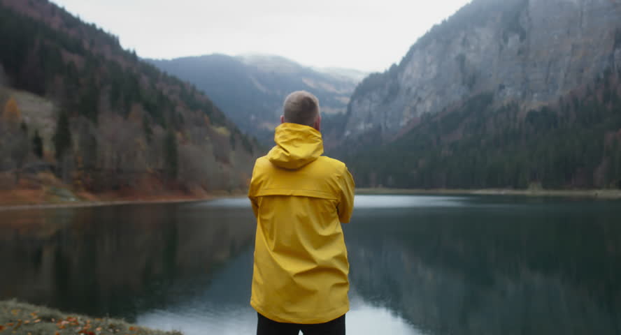 Back view of Caucasian male hiker in yellow raincoat stands near beautiful lake in Alps, rainy weather, morning shot. 4K UHD 60 FPS SLO MO