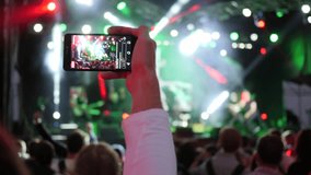 fan with smartphone into hand records video on background crowd in bright stage lights at rock festival in night time