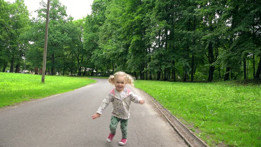 Active blond girl with pigtails running through park road in front of camera. Happy smiling child in summer. Handheld steadycam flycam movement front shot