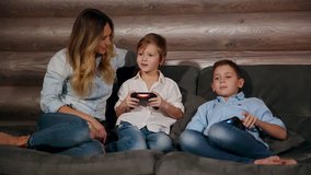 Mother and two sons sitting on the sofa in his house playing video games with wireless joystick. Happy people in the house.