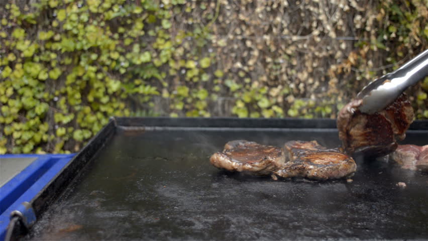Steaks being cooked on a BBQ, with a hand coming in to turn them over with tongs