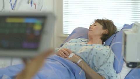 Camera rack focuses from a portable patient monitor held by a doctor to a mature woman lying in a hospital bed.