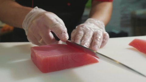 Close-up of sushi chef in gloves slices fresh fish at sushi bar. Professional cook cutting fish fillet at commercial kitchen, slow motion.