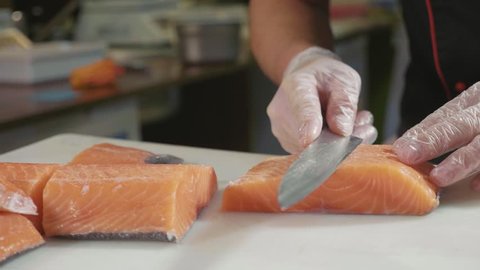 Close-up of sushi chef in gloves slices fresh salmon at sushi bar. Professional cook cutting fish fillet at commercial kitchen, slow motion.