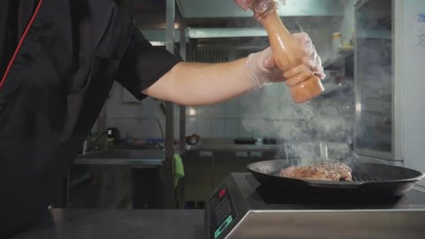 Meat frying on griddle pan. Crop of chef pouring spices in cooking steak, at restaurant commercial kitchen, slide slow motion