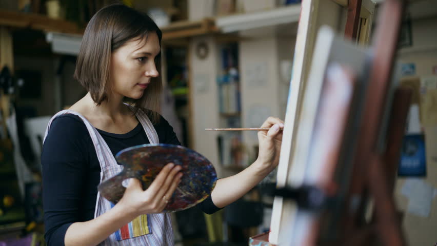 Young woman artist in apron painting picture on canvas in art studio