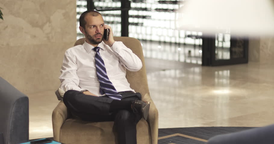 A stylish businessman uses a mobile phone sitting in the hotel lobby or office. A bearded man in a white shirt and tie is sitting in an armchair crossed legs. | Shutterstock HD Video #33294157