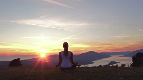 Young attractive girl sitting on the peak of a mountain. Practicing yoga. Surya Namaskar or Salutation to the Sun