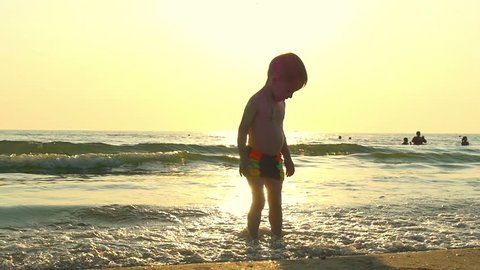 The child stands in the water on the sea beach, in the incoming waves, against the background of a decline at a slowed pace