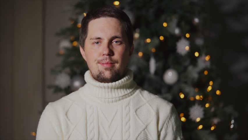 portrait of a young man in a winter sweater who stands near the Christmas tree, a Christmas tree decorated with toys and lights, calm people ready to celebrate with friends