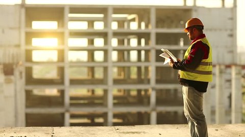 Master builder looks at new construction site and makes notes in works documentation. Worker in hard hat stands on project site and looks at new building structure, beautiful sunset at background