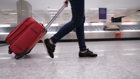 Passenger woman walk with trolley case against baggage carousel, low half view of slender female legs and medium wheeled bag, smooth tracking shot. Luggage conveyor belt number nine seen on background