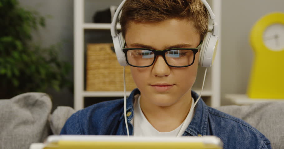 Teen boy in glasses and big white headphones working on his tablet computer in the living room. Close up. portrait. Indoors | Shutterstock HD Video #33316525