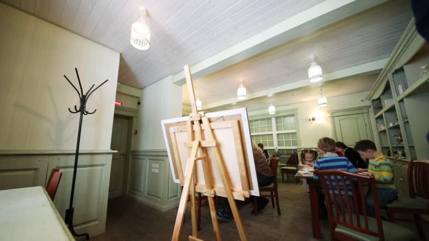 Five children draws sitting at tables in a room with a canvas on an easel