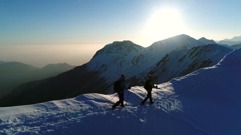 Aerial - Flyover two adult male mountaineers hiking on snowy mountain top at winter sunset. Men equipped with warm clothes, trekking poles and backpacks walking on mountain peak in Julian Alps : vidéo de stock