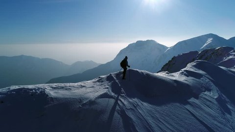 Aerial, slow motion - Young man hiking on top of snowy mountain at beautiful winter sunset. Male mountaineer with trekking poles and a backpack walking on mountain ridge in Julian Alps