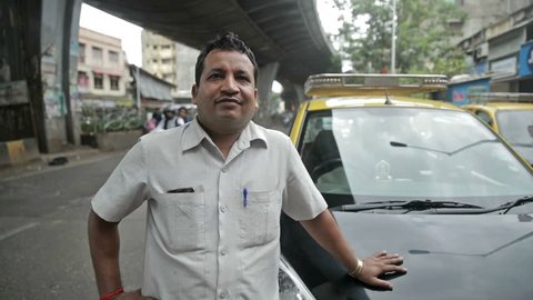 A proud and smiling taxi driver standing in front of his taxi while vehicles passing in the background. Real people of India
