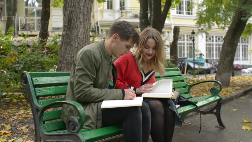 Guy and Girl Students on a Bench in a Park write an Abstract read a Textbook