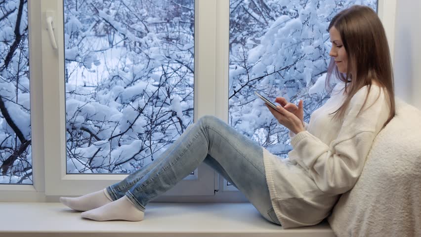 Pretty girl sitting on the windowsill, looking the window and using tablet computer. Winter outside.