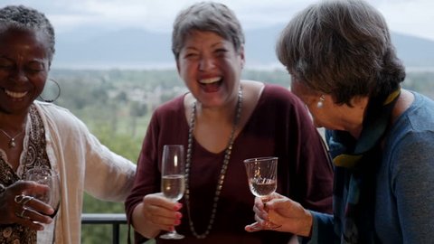 Mixed racial group of senior women friends in their 60s laughing out loud on a balcony holding wine glasses with an amazing mountain view in slow motion