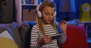 Cute blonde girl in the big white headphones watching music video on her smart phone and dancing in the rhythm of music in the living room late at night. indoors