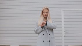 A young fashionably dressed blonde girl in a coat uses a smart bracelet standing outdoors. Slow motion video